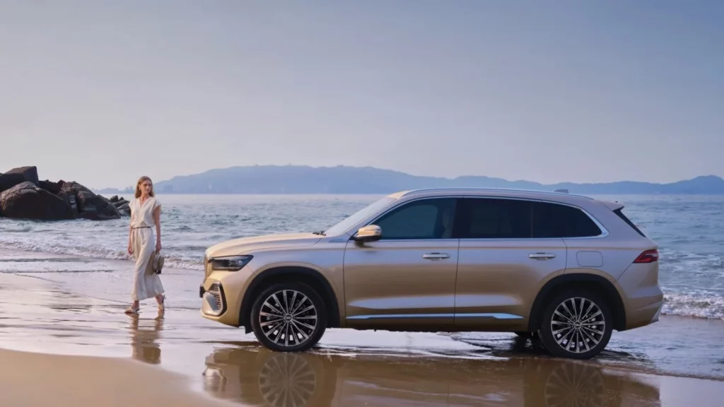 Złoty SUV Geely Monjaro stoi na piaszczystej plaży obok spacerującej kobiety